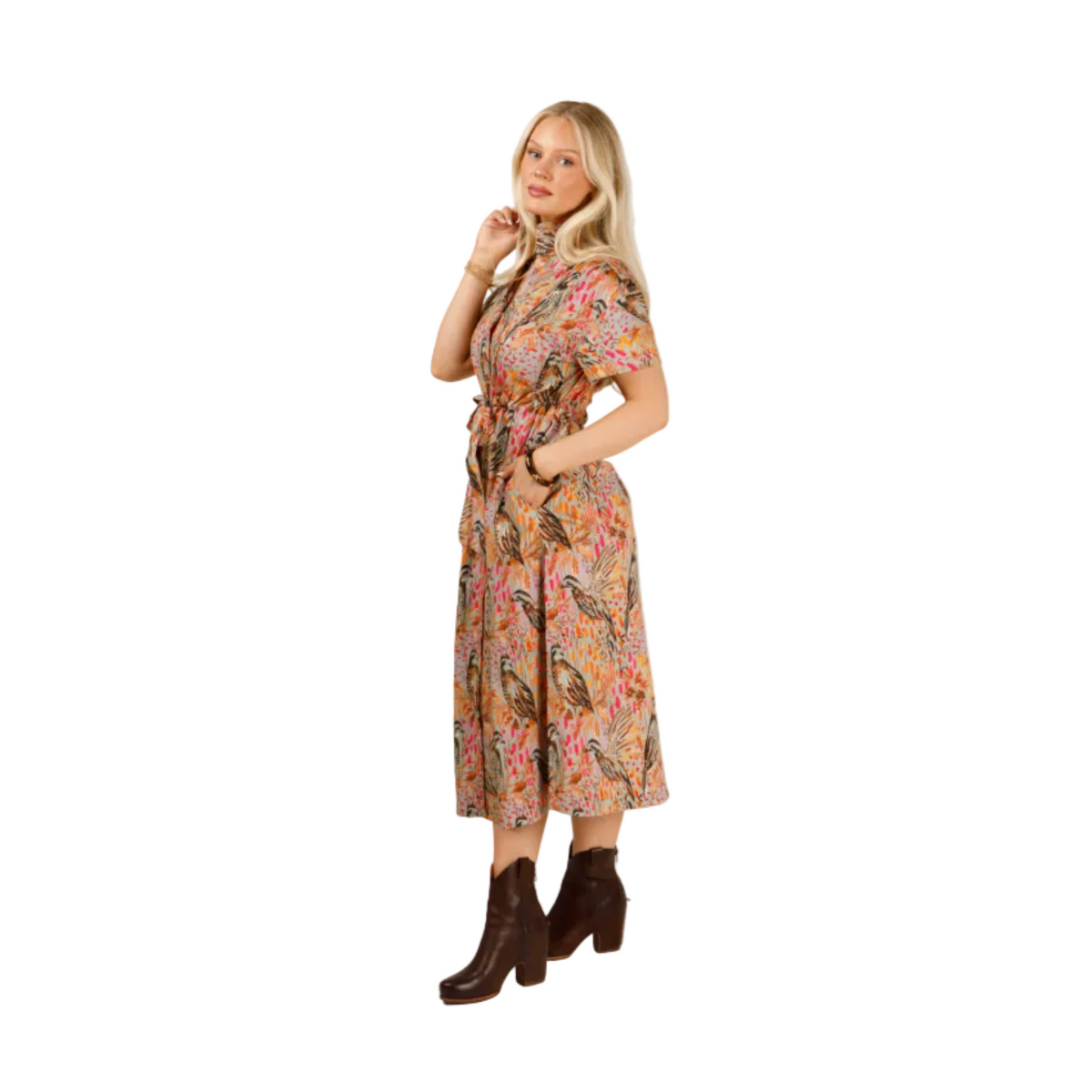 A woman modeling a short-sleeved midi dress with a colorful paisley pattern, paired with dark brown ankle boots, against a plain light gray studio background.