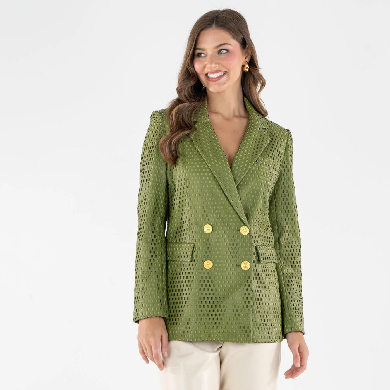 A woman modeling a sage green double-breasted blazer with a textured, perforated pattern and gold buttons, paired with cream trousers, against a plain white studio background.