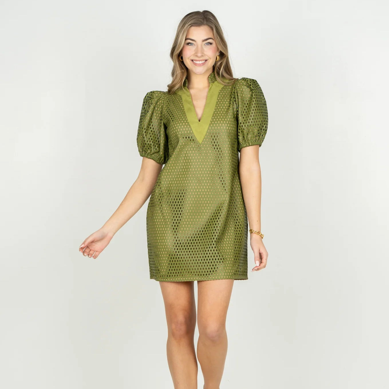 A woman modeling a short olive green puff-sleeve dress with a textured, dotted mesh-like pattern and a V-neckline, set against a plain white studio background.