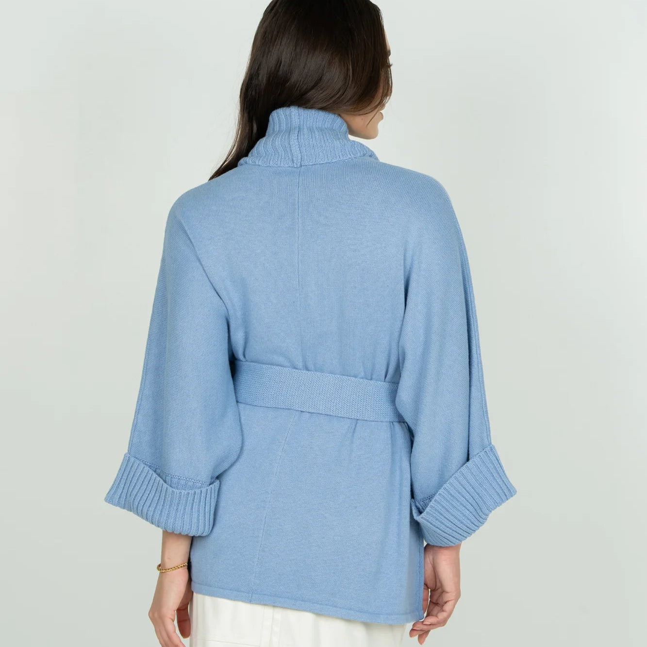 A woman shown from the back wearing a light blue knit wrap-style cardigan with a high mock neck, waist tie, and ribbed cuffs, paired with white pants against a light gray studio background.