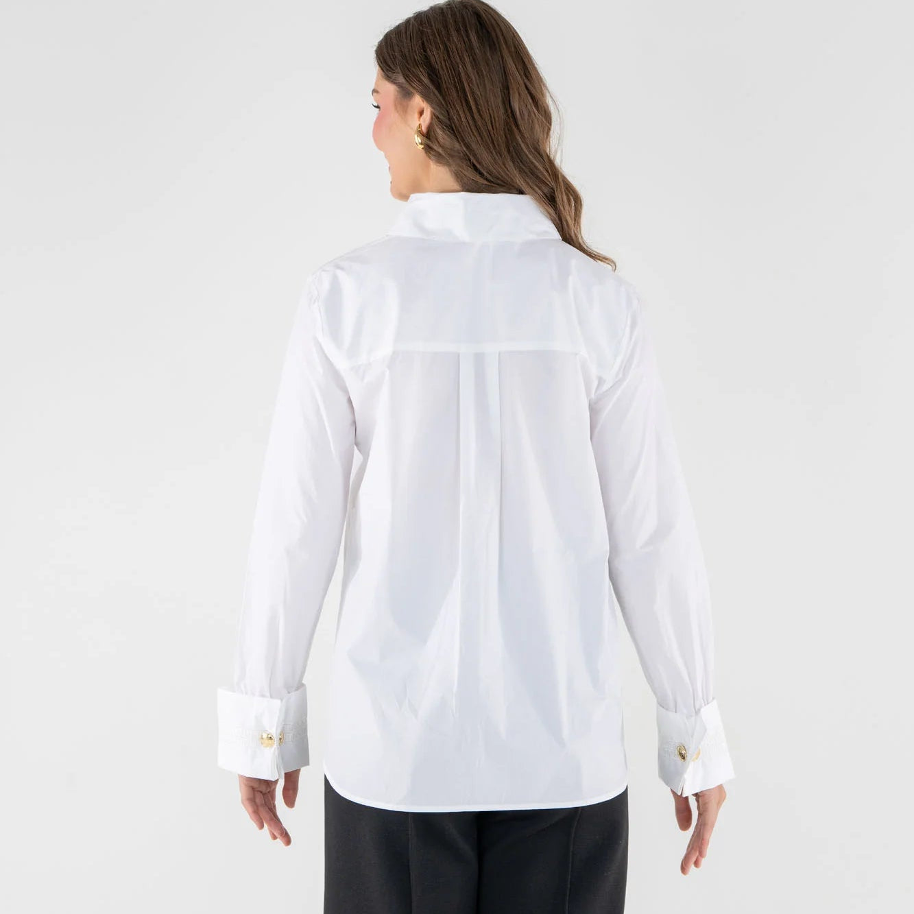 A woman shown from the back wearing a white long-sleeve button-up shirt with a high collar and dark wide-leg trousers, photographed against a light gray studio background.