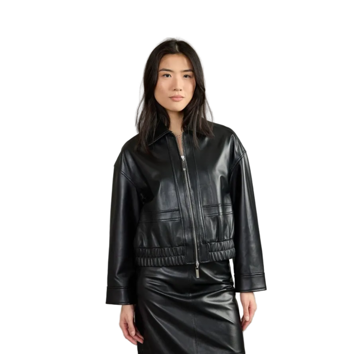 A model wearing a black leather cropped jacket with a front zipper and a matching black leather skirt, photographed against a solid gray studio background.