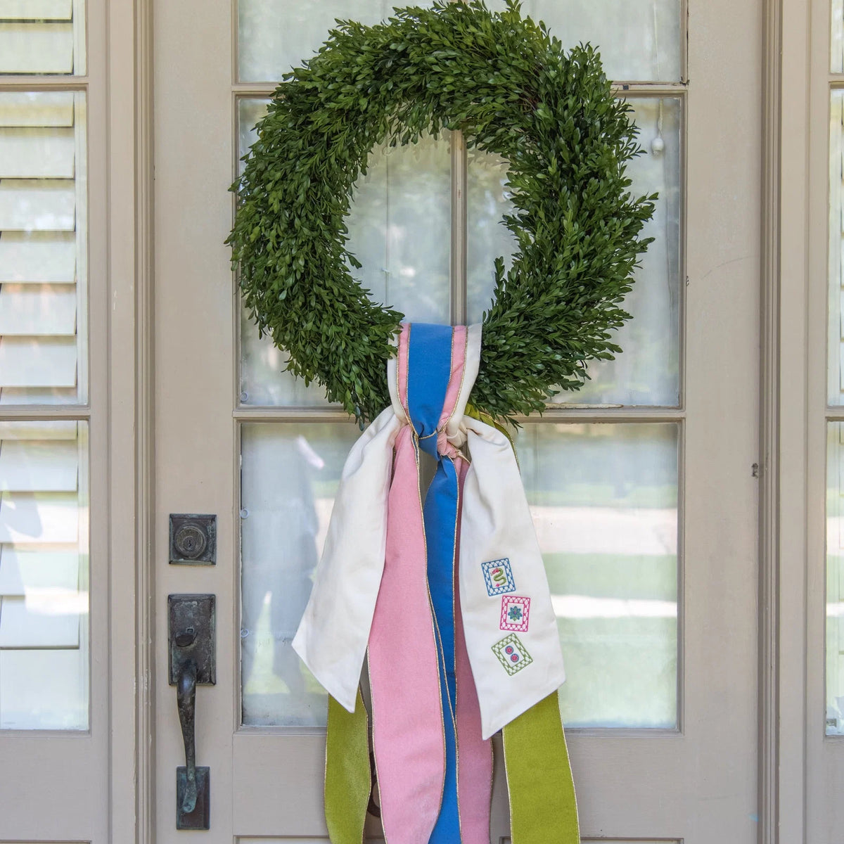 A green wreath hanging on a front door, decorated with long hanging ribbons in white, pink, blue, and olive green.