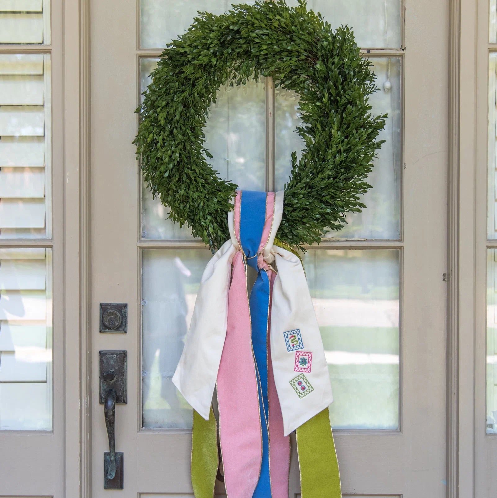 A green wreath hanging on a front door, decorated with long hanging ribbons in white, pink, blue, and olive green.