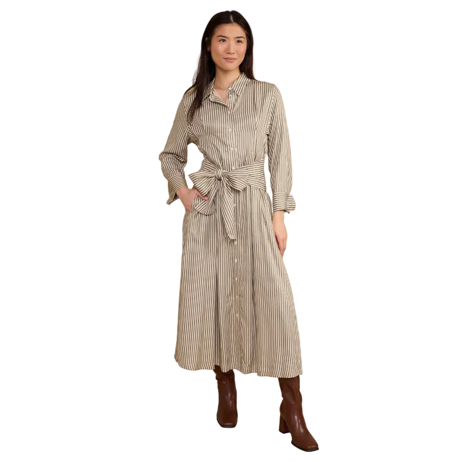 A woman modeling a long-sleeve button-front shirt dress with thin vertical stripes in beige and brown, featuring a tie waist belt and brown ankle boots, against a solid tan studio background.