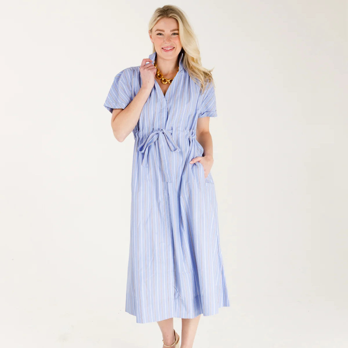 A woman modeling a light blue and white vertically striped midi dress with short puff sleeves, a tie waist, and side pockets, paired with gold sandals against a plain white studio background.