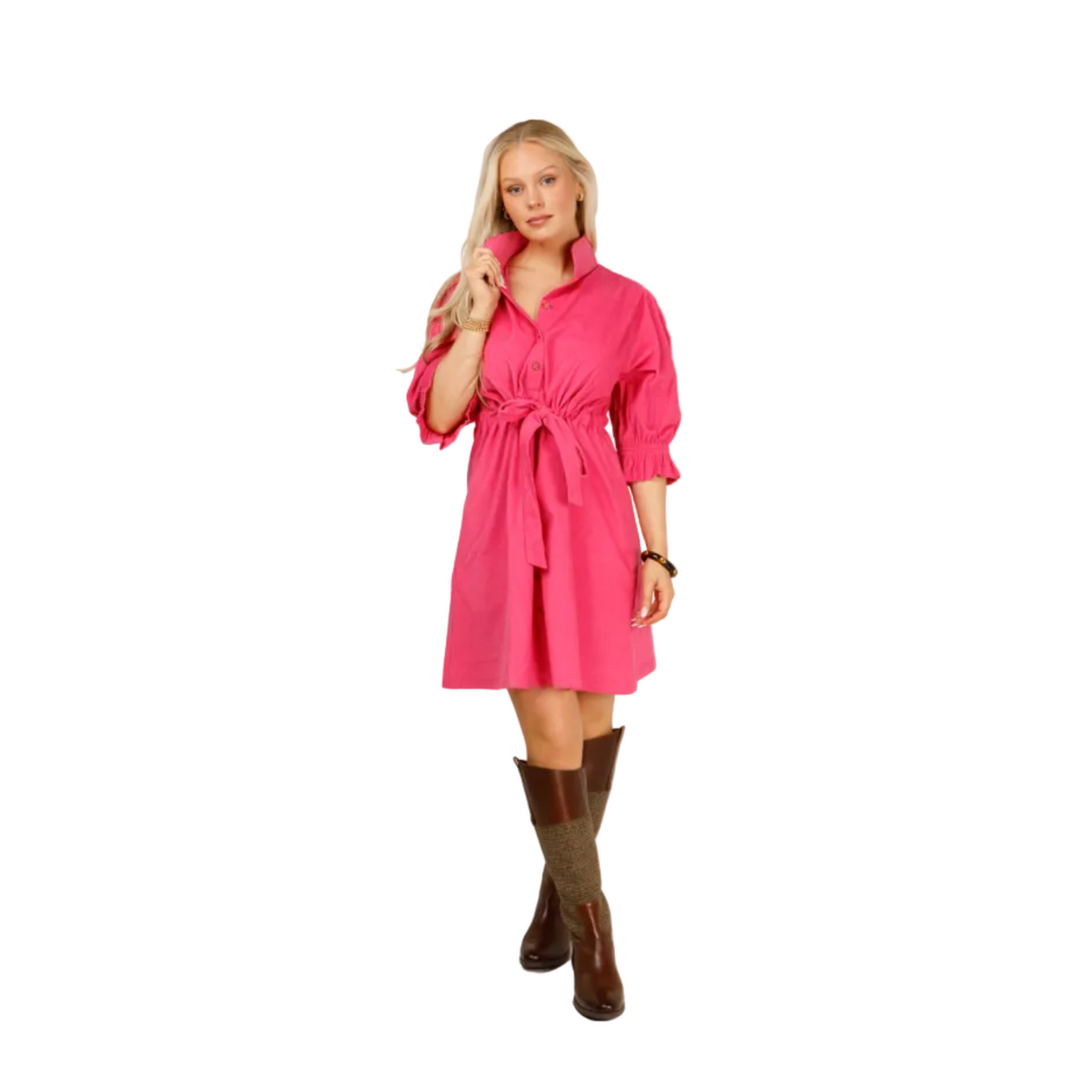 A woman modeling a bright pink collared shirt dress with a tie waist and puffed sleeves, paired with brown knee-high boots, against a light gray studio background.
