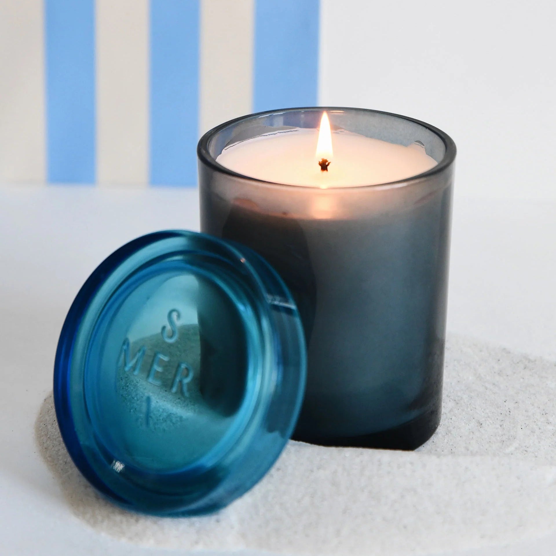 A lit candle in a dark smoky glass jar sits beside a matching blue translucent lid embossed with 'SMEER' on a white textured surface, with blue and white vertical stripes in the background.
