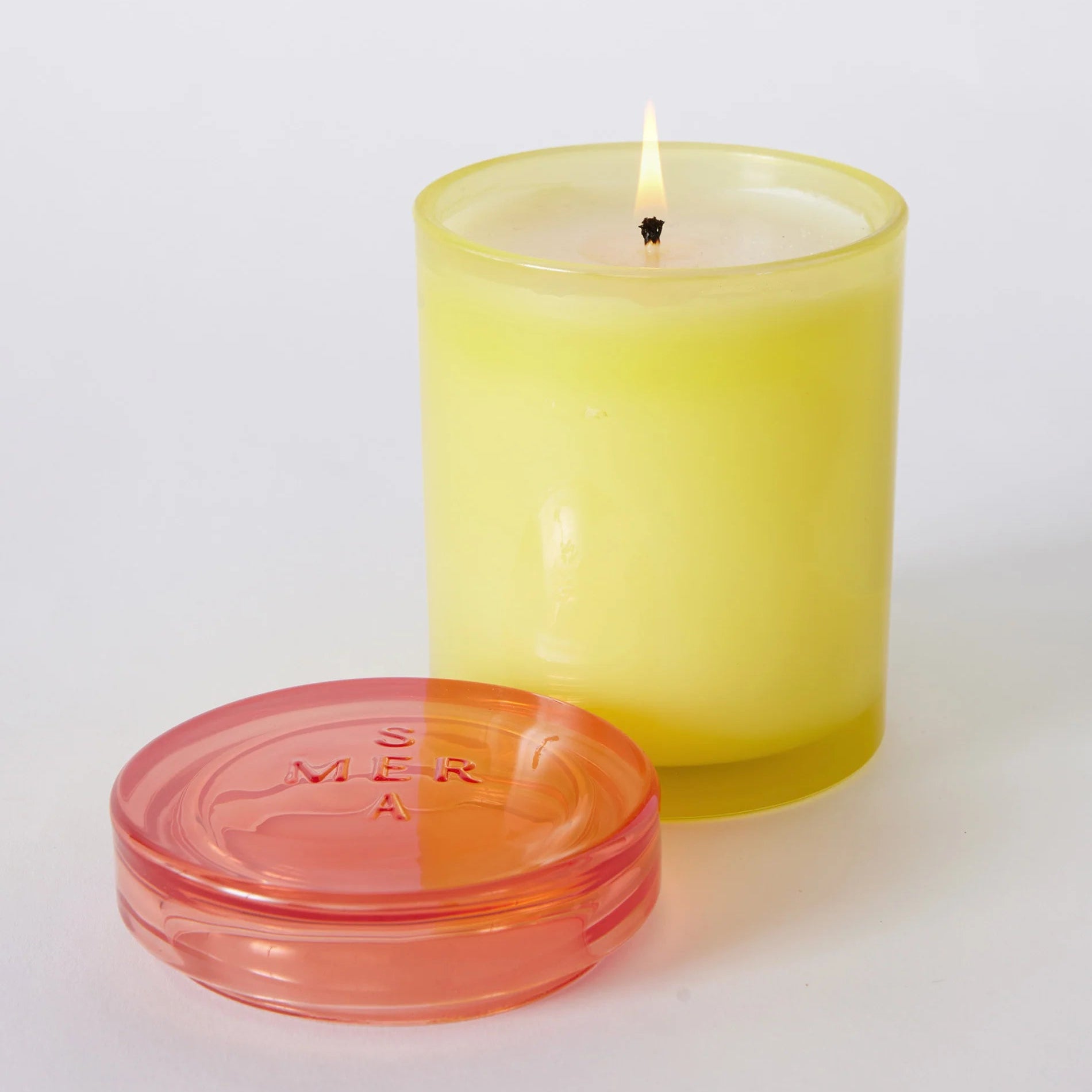 A lit yellow candle in a translucent yellow glass holder with a separate translucent pink lid placed in front, photographed on a clean white background.
