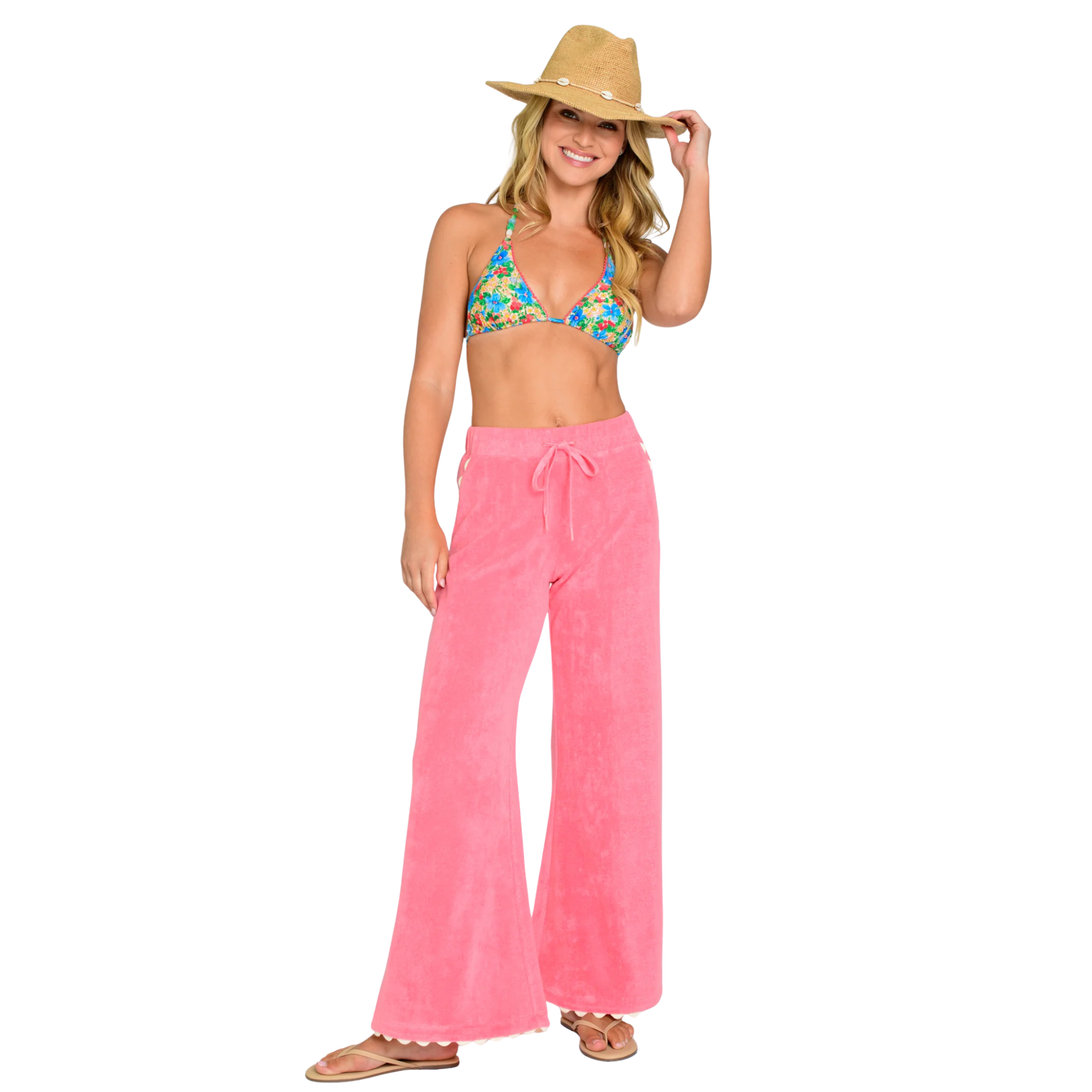 Woman wearing a floral bikini top and pink pants with a straw hat on a white background