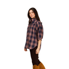 A woman modeling a high-neck, three-quarter sleeve plaid button-up shirt in navy and dusty pink, paired with dark brown pants and tan suede knee-high boots, against a plain light gray studio background.