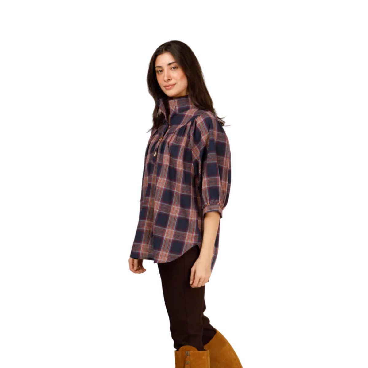 A woman modeling a high-neck, three-quarter sleeve plaid button-up shirt in navy and dusty pink, paired with dark brown pants and tan suede knee-high boots, against a plain light gray studio background.