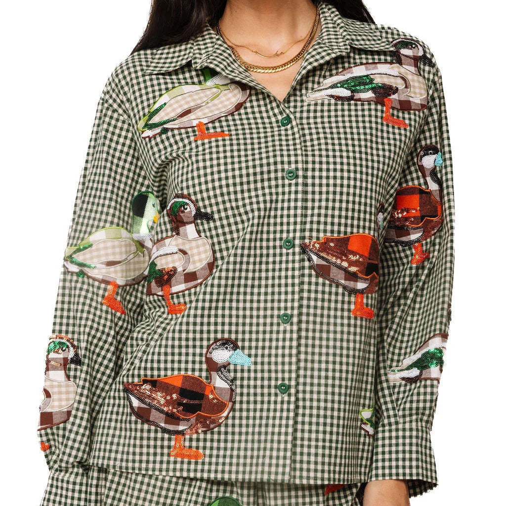 A woman wearing a matching long-sleeve button-up shirt and shorts pajama set in a green and white gingham pattern, decorated with realistic embroidered ducks in various colors.