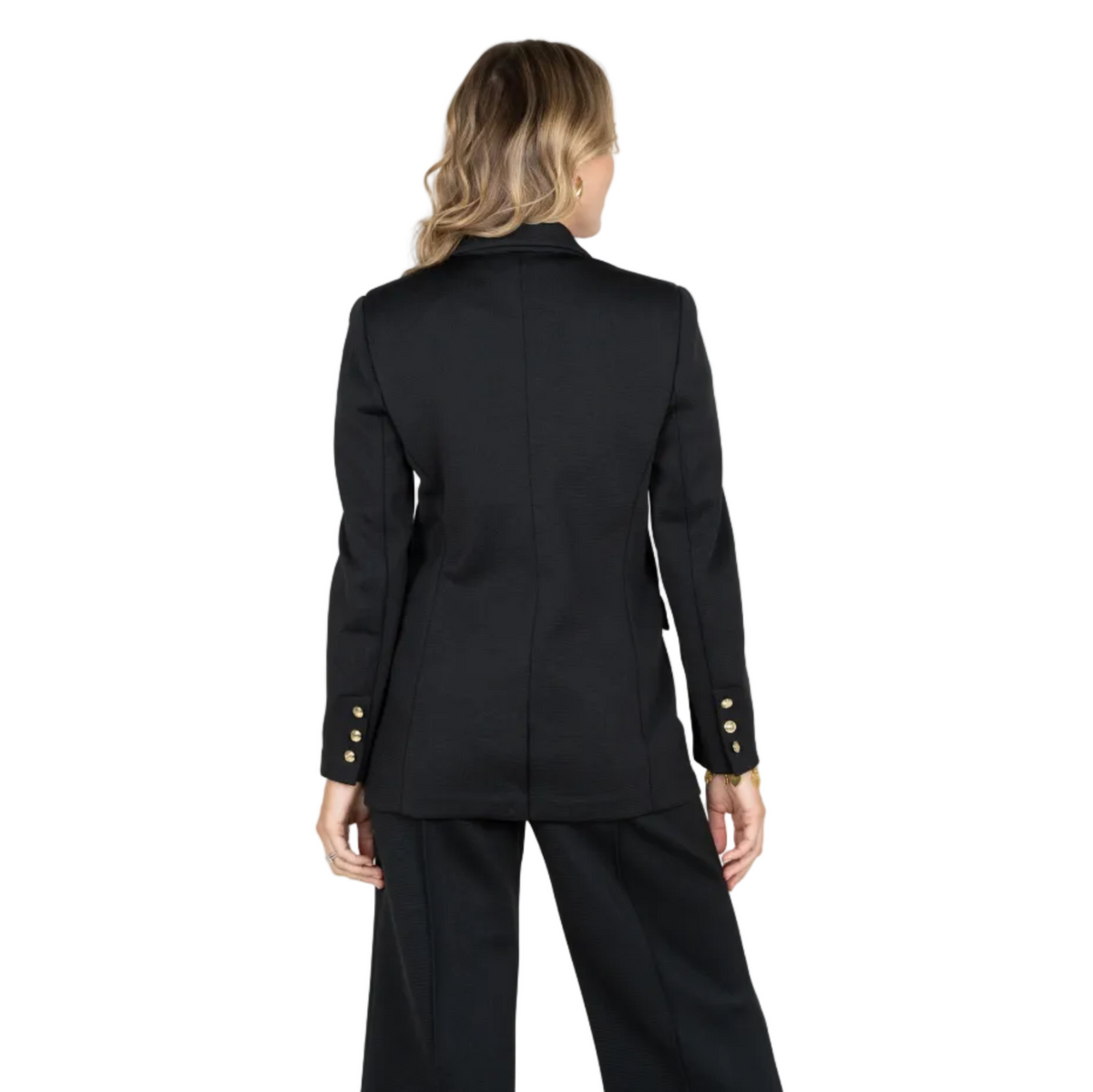 A woman shown from the back wearing a black tailored blazer with gold button accents on the sleeves and matching black trousers, photographed against a light gray studio background.