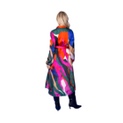 A woman stands with her back to the camera wearing a long-sleeved, belted trench coat with a vibrant multicolor abstract print, set against a solid blue studio background.