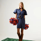 A woman stands on a football field yard-line mat wearing a navy blue short-sleeve shirt dress with a gold buckle belt, brown knee-high boots, and holding red and blue pom-poms. A brown football is on the mat in front of her against a light gray studio background.