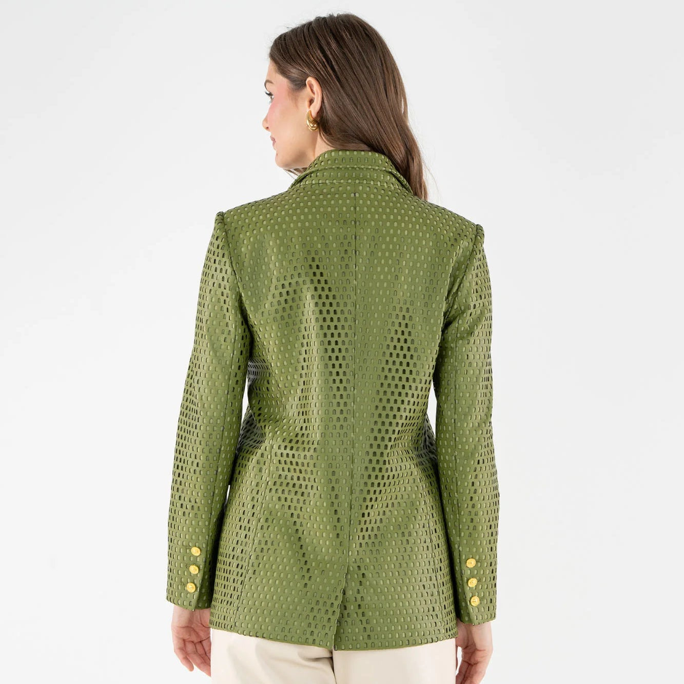 A woman viewed from the back wearing an olive green textured leather blazer with a geometric pattern and gold buttons, paired with cream pants, against a plain white studio background.