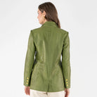 A woman viewed from the back wearing an olive green textured leather blazer with a geometric pattern and gold buttons, paired with cream pants, against a plain white studio background.