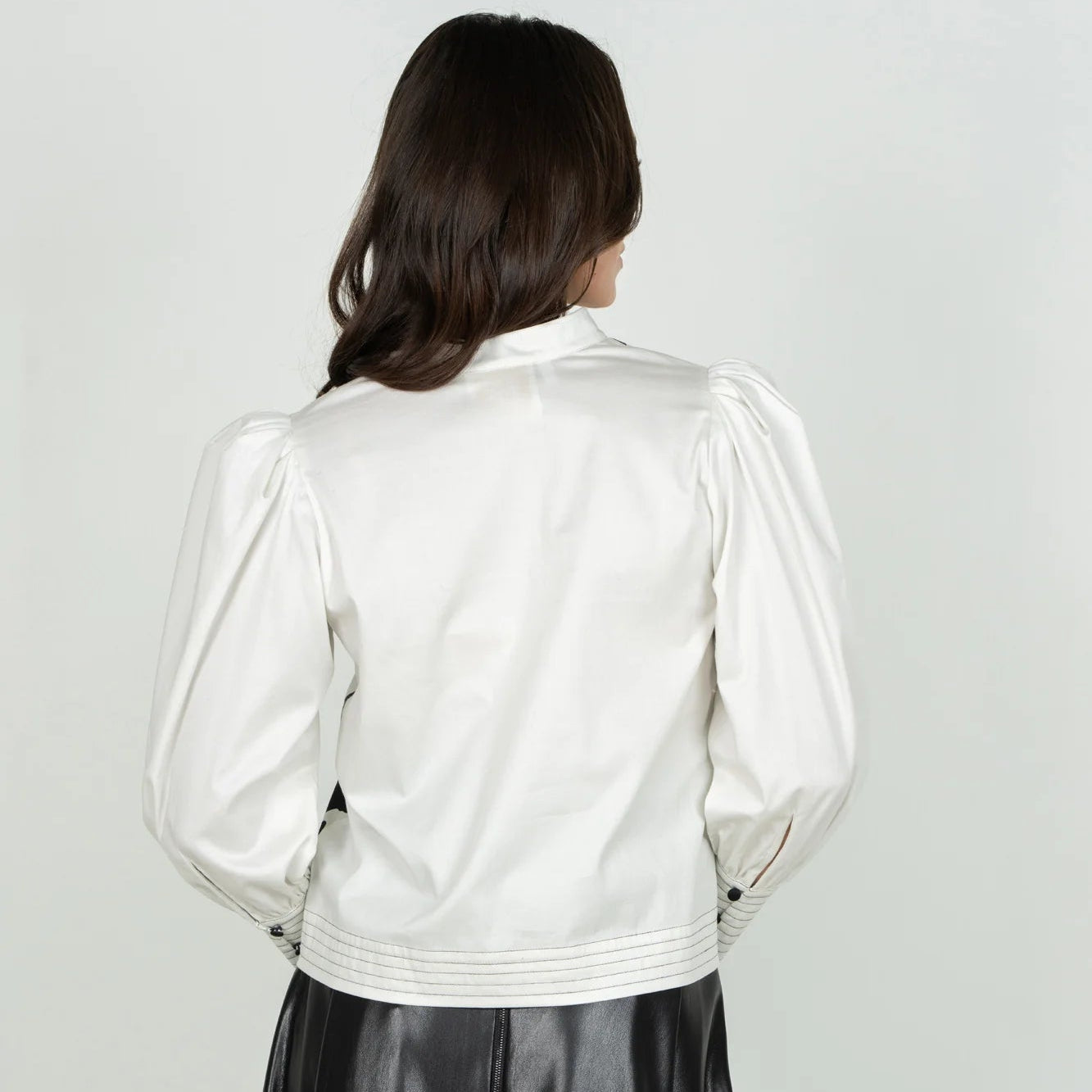A woman shown from the back wearing a white cropped jacket with puffed long sleeves and a black skirt, against a plain light gray studio background.
