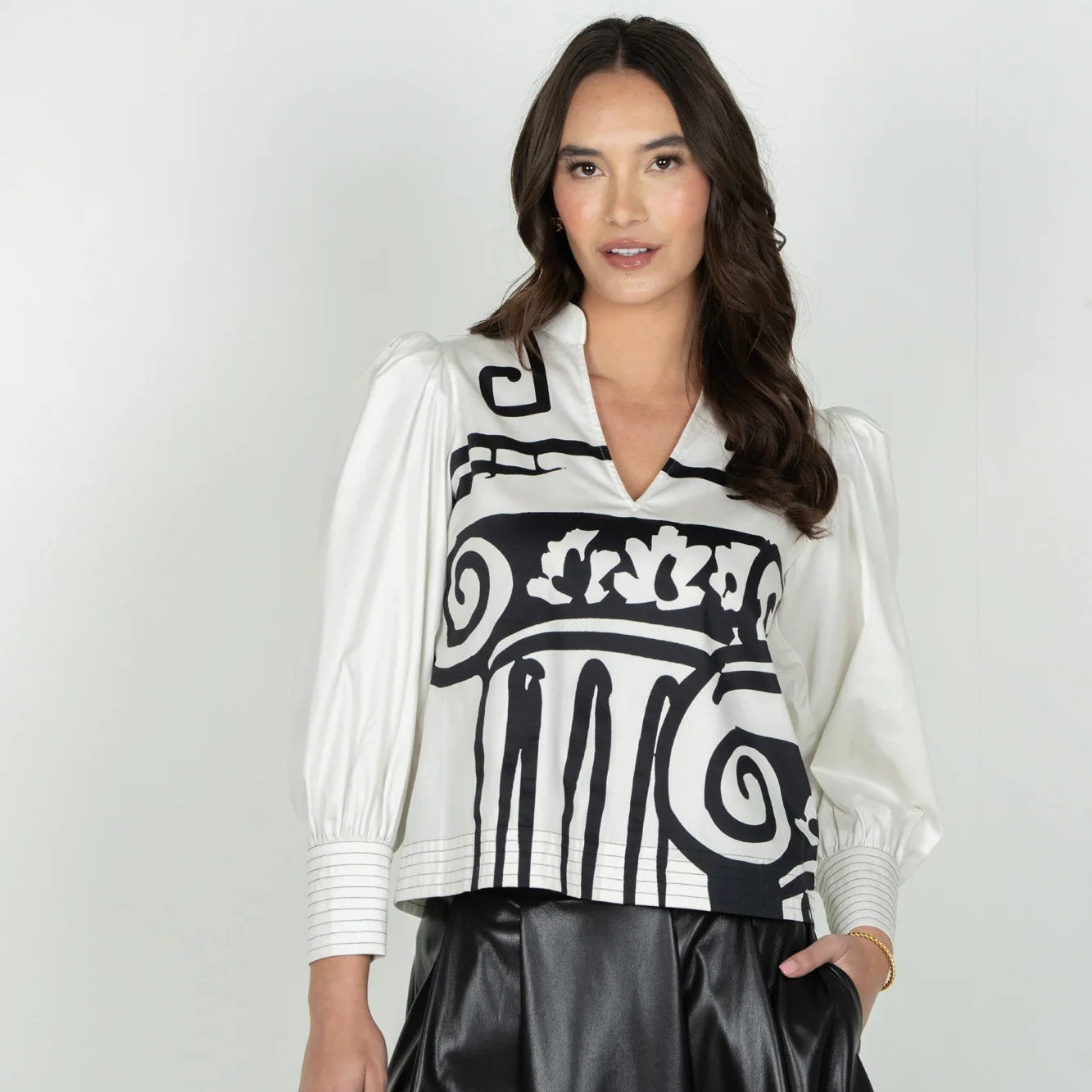 A woman modeling a white long-sleeve blouse with a bold black classical column pattern on the front, paired with a black satin skirt, against a light gray studio background.