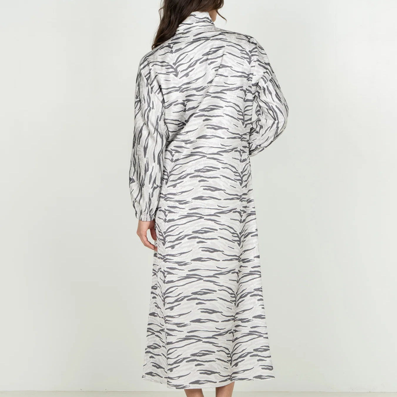 A woman viewed from behind wearing a long-sleeved collared shirt dress with a black and white zebra-like abstract pattern, paired with black flat sandals, against a plain white studio background.