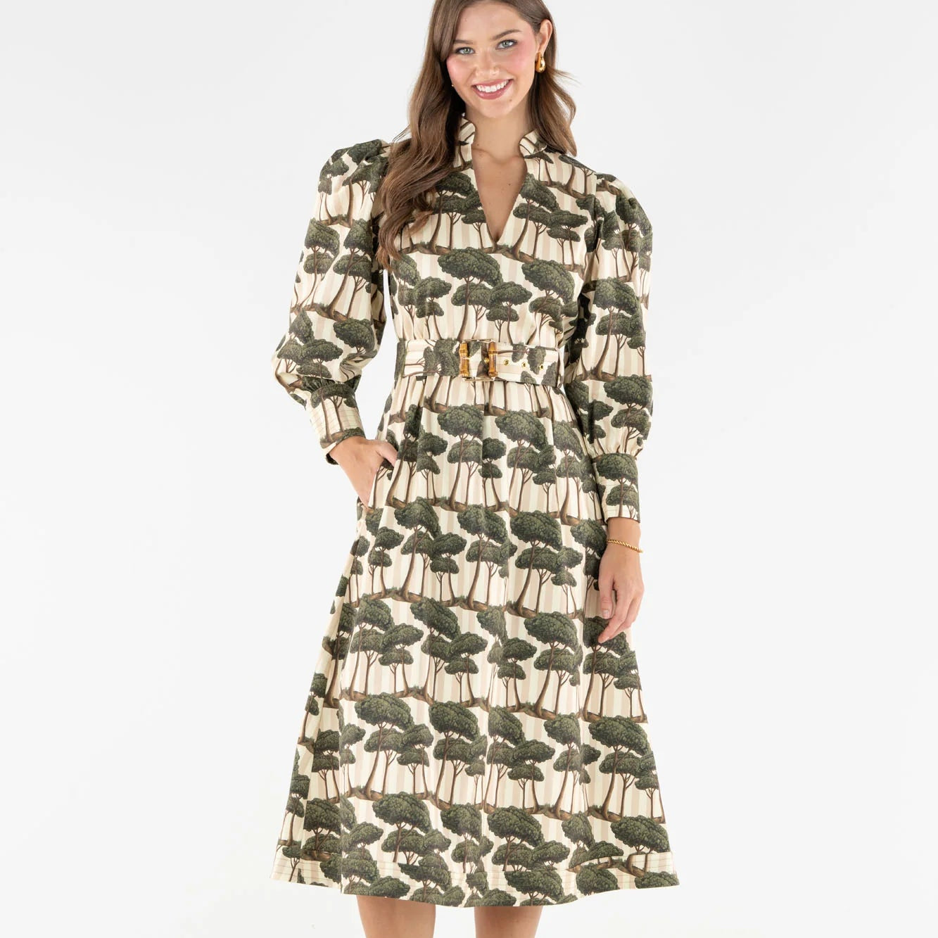 A woman modeling a long-sleeved midi dress with a tree print pattern in olive green and beige, featuring a belted waist and collar, set against a plain white studio background.