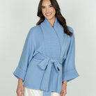 A woman wearing a light blue ribbed knit wrap robe with a tie belt and wide three-quarter sleeves, paired with white pants, against a light gray studio background.