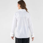 A woman shown from the back wearing a white long-sleeve button-up shirt with a high collar and dark wide-leg trousers, photographed against a light gray studio background.