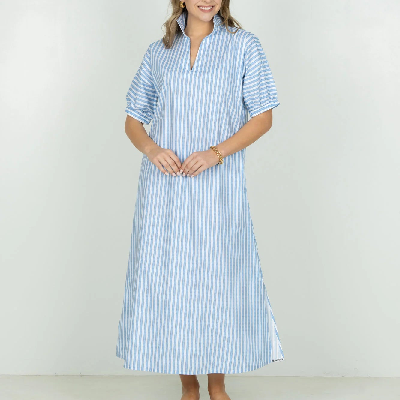 A woman modeling a light blue and white vertically striped midi dress with short sleeves and a split neckline, standing against a plain white studio background.
