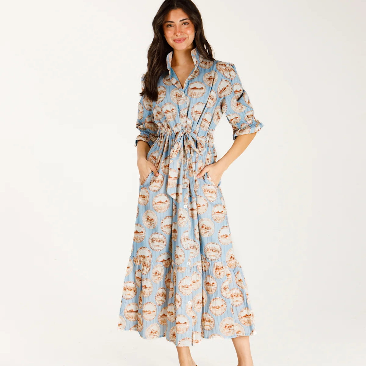 A woman modeling a light blue midi shirt dress with a brown and beige coffee cup pattern, featuring a tie waist and pockets, paired with brown slide sandals against a plain white studio background.