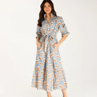 A woman modeling a light blue midi shirt dress with a brown and beige coffee cup pattern, featuring a tie waist and pockets, paired with brown slide sandals against a plain white studio background.
