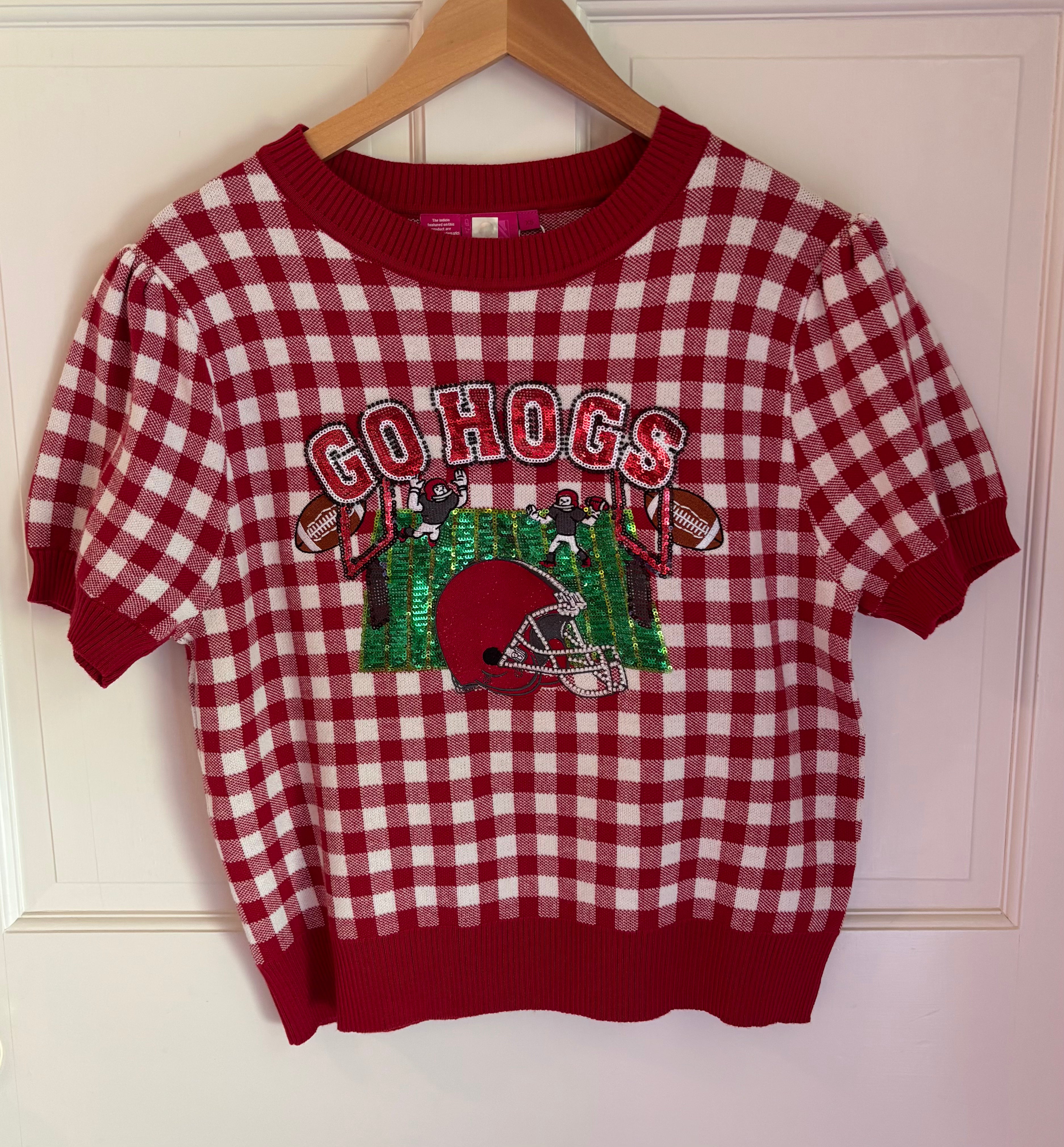 A red and white gingham knit crewneck sweater with a 'GO HOGS' football graphic on the front, hanging on a wooden hanger against a white paneled door.