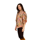 A woman modeling a colorful, patterned puff-sleeve blouse with dark brown cropped pants and tan suede knee-high boots, standing against a plain light gray studio background.