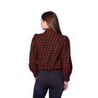 A woman shown from the back wearing a black blouse with a repeating red and orange abstract pattern, paired with dark pants, set against a solid blue studio background.