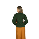 A woman shown from the back wearing a dark green knit sweater with brown leather-like trim details and a matching rust-orange skirt, photographed against a plain light gray studio background.
