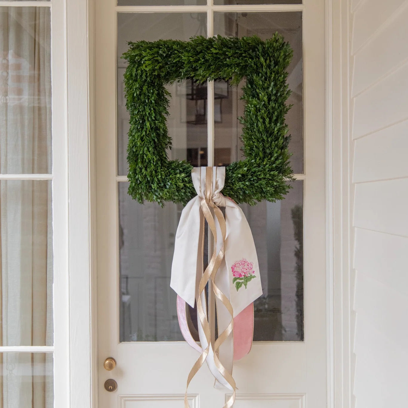 A square green boxwood wreath hanging on a white front door with a large cream ribbon and pink fabric accents tied around it.