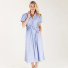 A woman modeling a light blue and white vertically striped midi dress with short puff sleeves, a tie waist, and side pockets, paired with gold sandals against a plain white studio background.