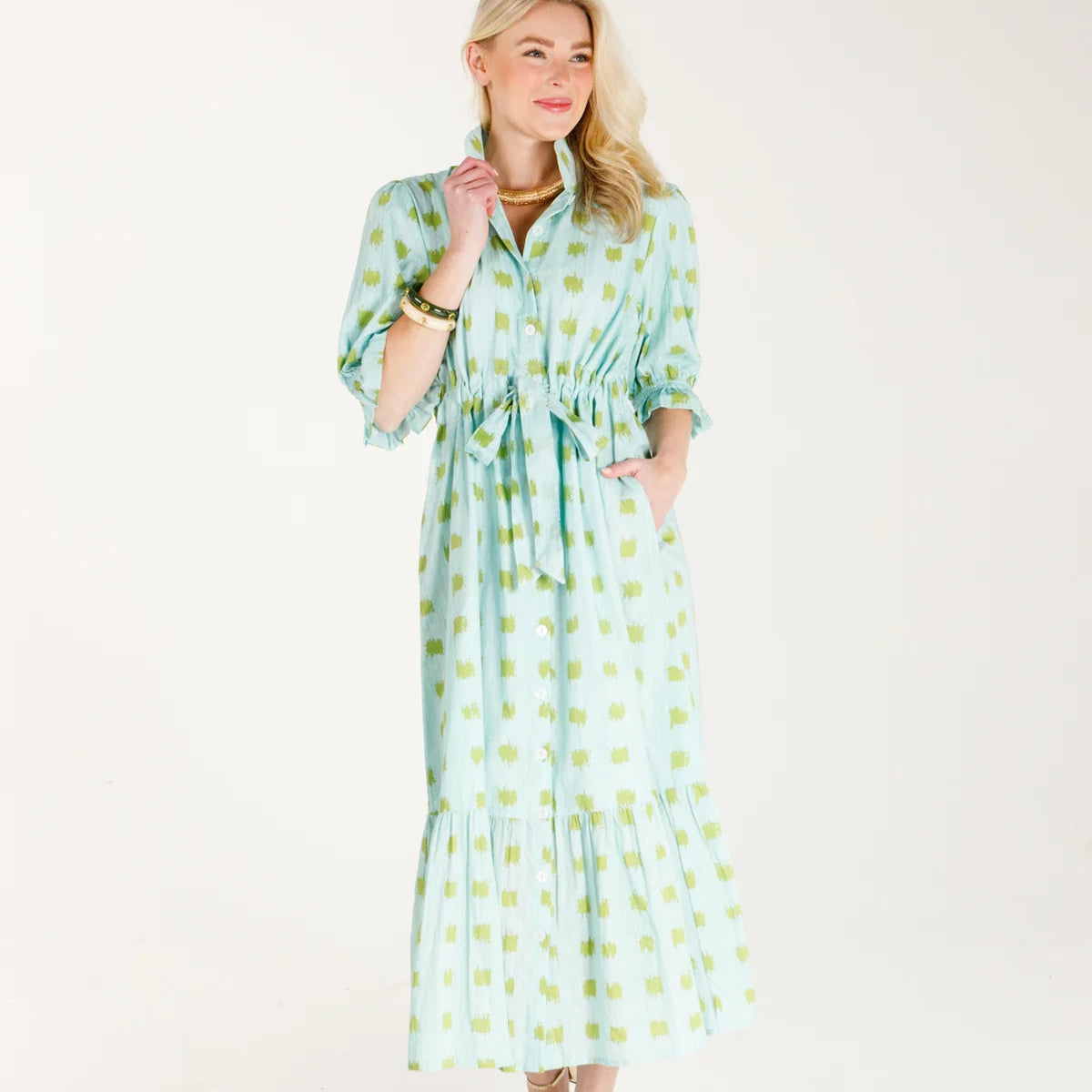A woman modeling a light green and white patterned midi dress with puff sleeves, a tie waist, and a ruffled hem, standing against a plain white studio background.