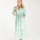 A woman modeling a light green and white patterned midi dress with puff sleeves, a tie waist, and a ruffled hem, standing against a plain white studio background.