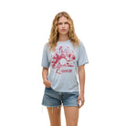A woman modeling a light blue graphic t-shirt with a red and white retro design and the word 'Queen' printed on the front, paired with denim shorts and black flats, against a solid beige background.