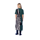 A woman stands with her back to the camera wearing a short-sleeve green dress with a large black and white zebra print panel on the back, paired with black boots, set against a solid blue studio background.