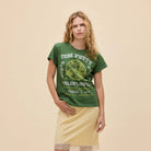 A woman modeling a green graphic t-shirt with 'TOM FETTS' text and a pale yellow skirt, standing against a solid beige background.