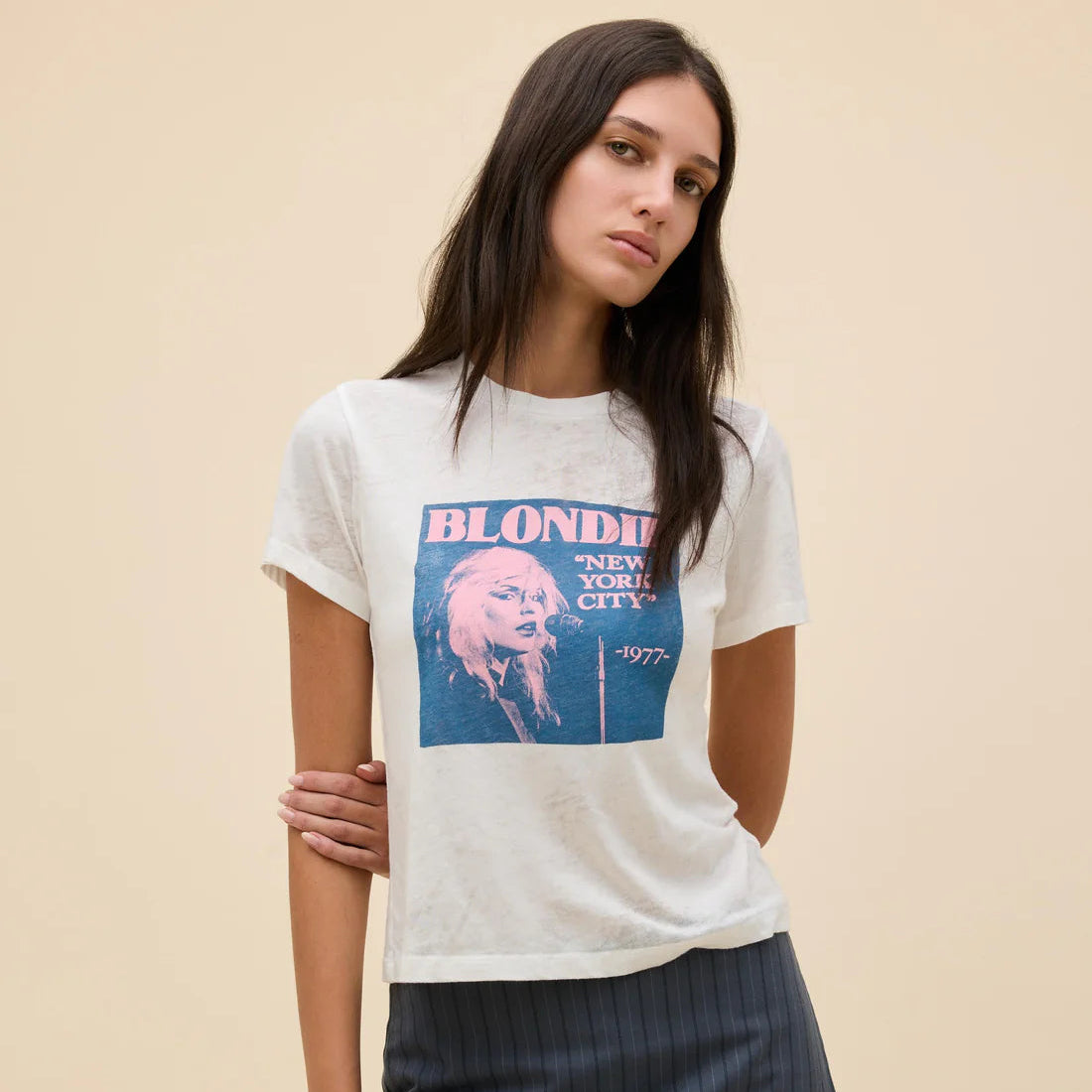 A woman modeling a white short-sleeve graphic T-shirt with a blue and pink 'Blondie' print, paired with dark pinstriped pants, against a solid beige background.