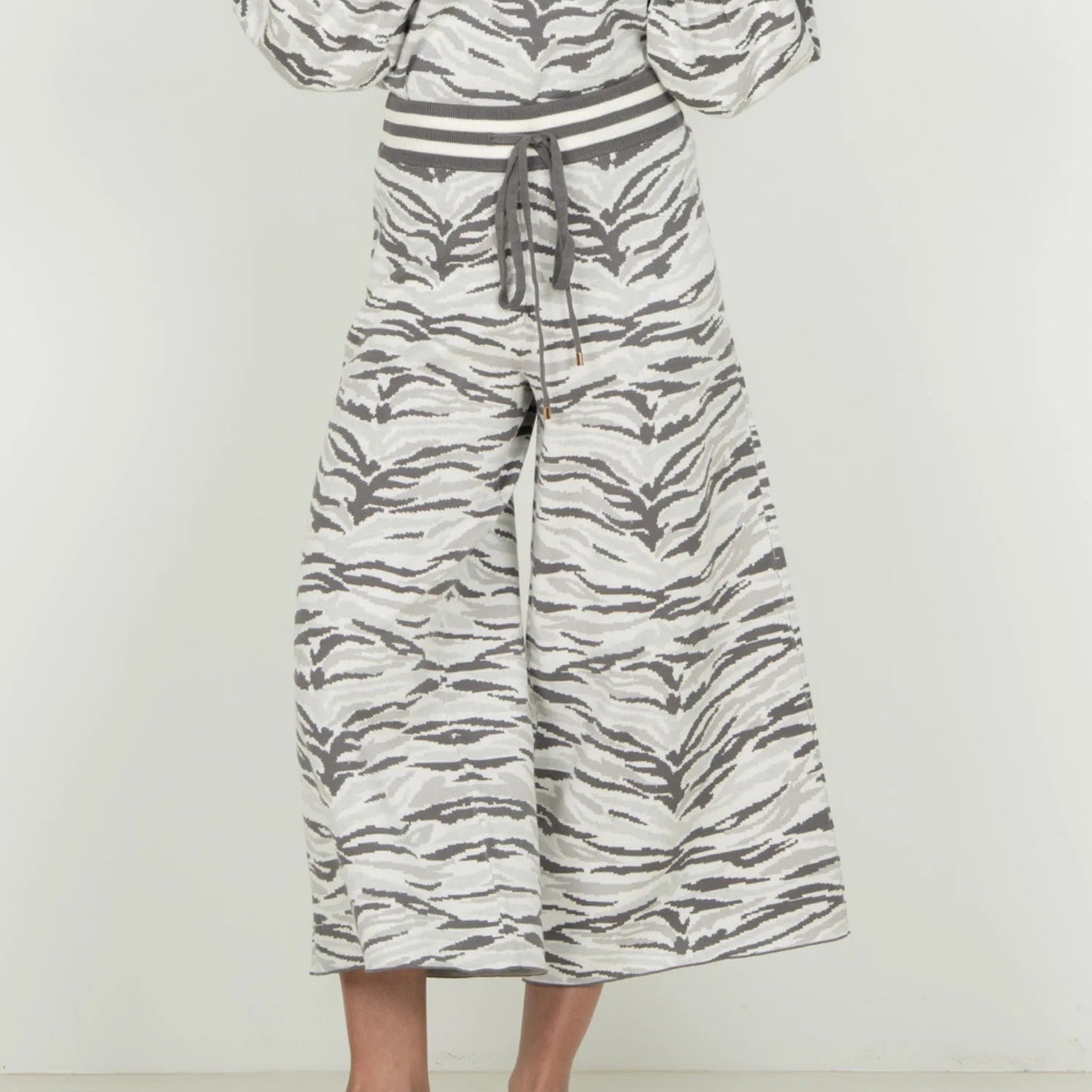 A model is shown from the back wearing a matching two-piece set consisting of a long-sleeve top and wide-leg cropped pants, both featuring a gray and white tiger-stripe pattern. The outfit is styled with light beige flat loafers against a plain white studio background.