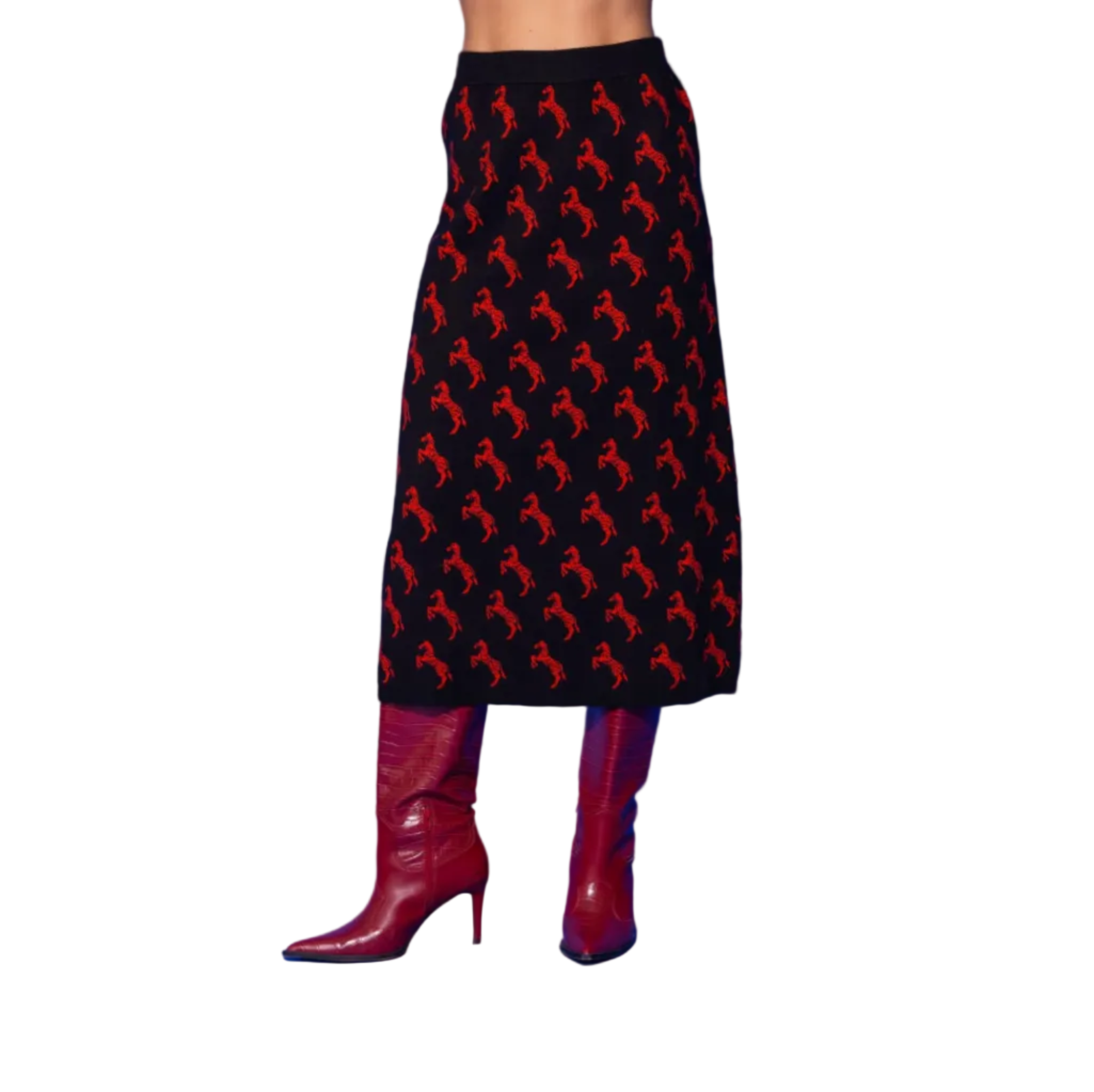 A model from the waist down wears a black midi skirt with a repeating red horse pattern, paired with red leather knee-high boots, set against a solid blue studio background.