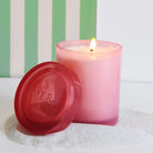 Pink candle with a red lid on a white surface with a striped green and white background